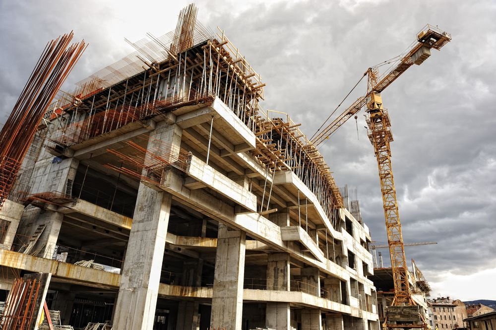 Construction site with a multi-story concrete building under construction, featuring a crane and cloudy sky.