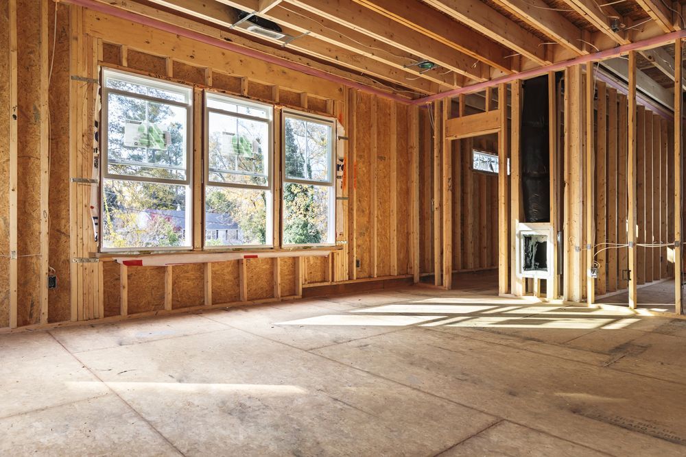 Interior of a house under construction, showing wooden framing, a window, and an open doorway. Sunlight streams in.