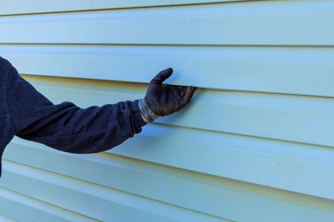 Gloved hand pulling on light blue vinyl siding on a house.