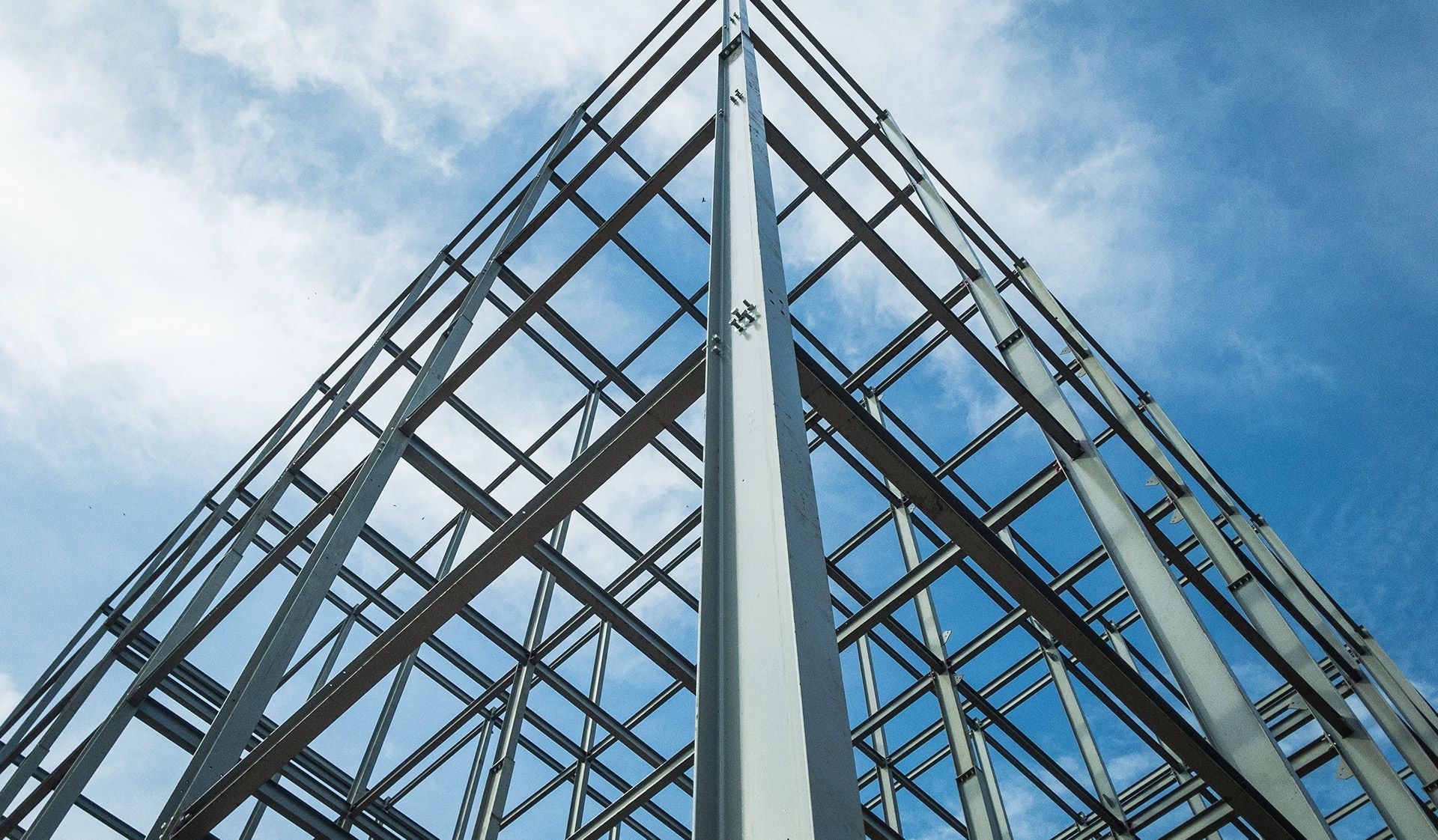 Steel framework of a building against a blue sky with white clouds. The perspective is upward, emphasizing the structure's grid pattern.