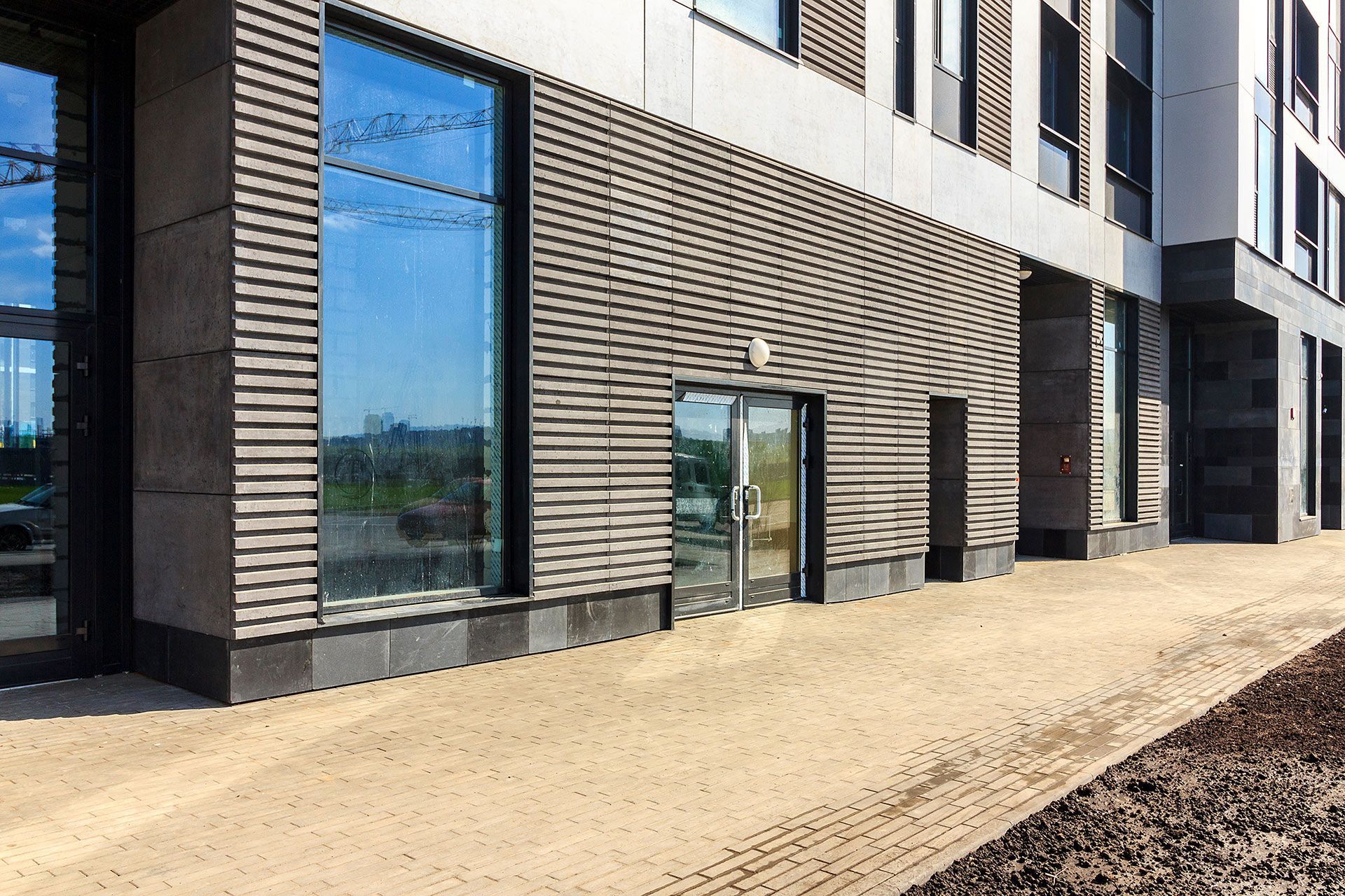 Exterior of a modern building with a glass door, large windows, and a paved walkway. The facade has gray paneling and dark accents.