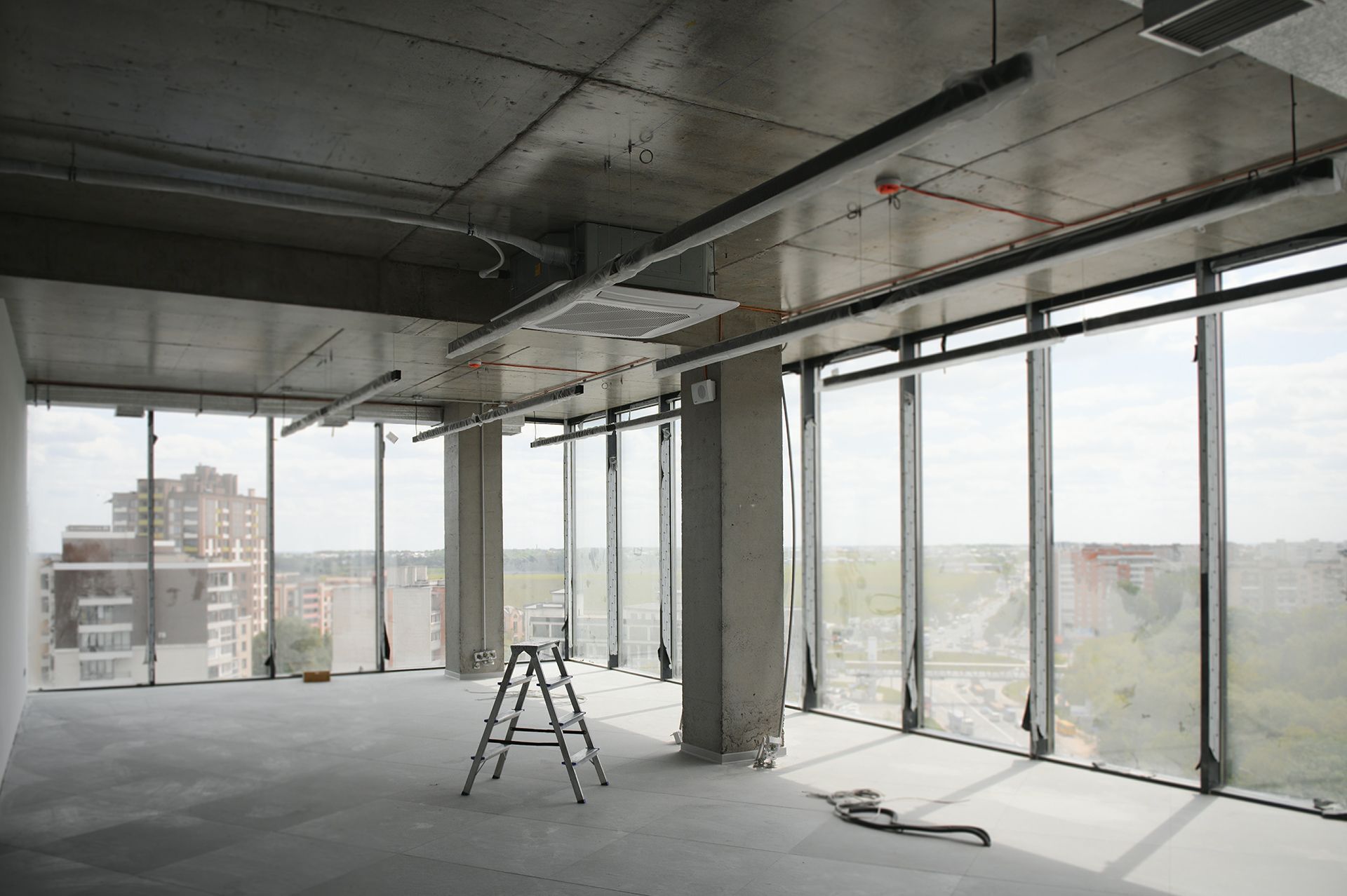 Interior view of an unfinished office space with large windows, a ladder, and exposed concrete surfaces.