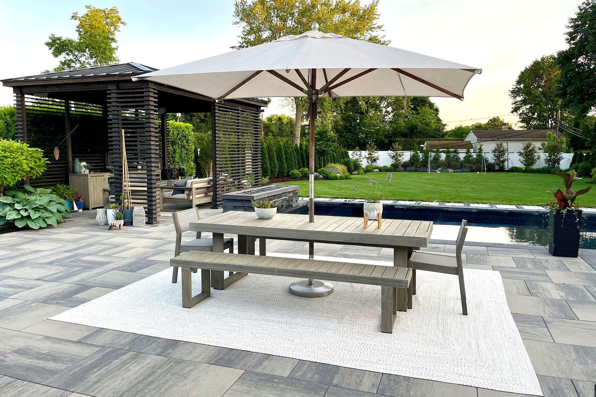 Outdoor patio with a wooden table and bench, surrounded by chairs, a pool, and a shaded structure.