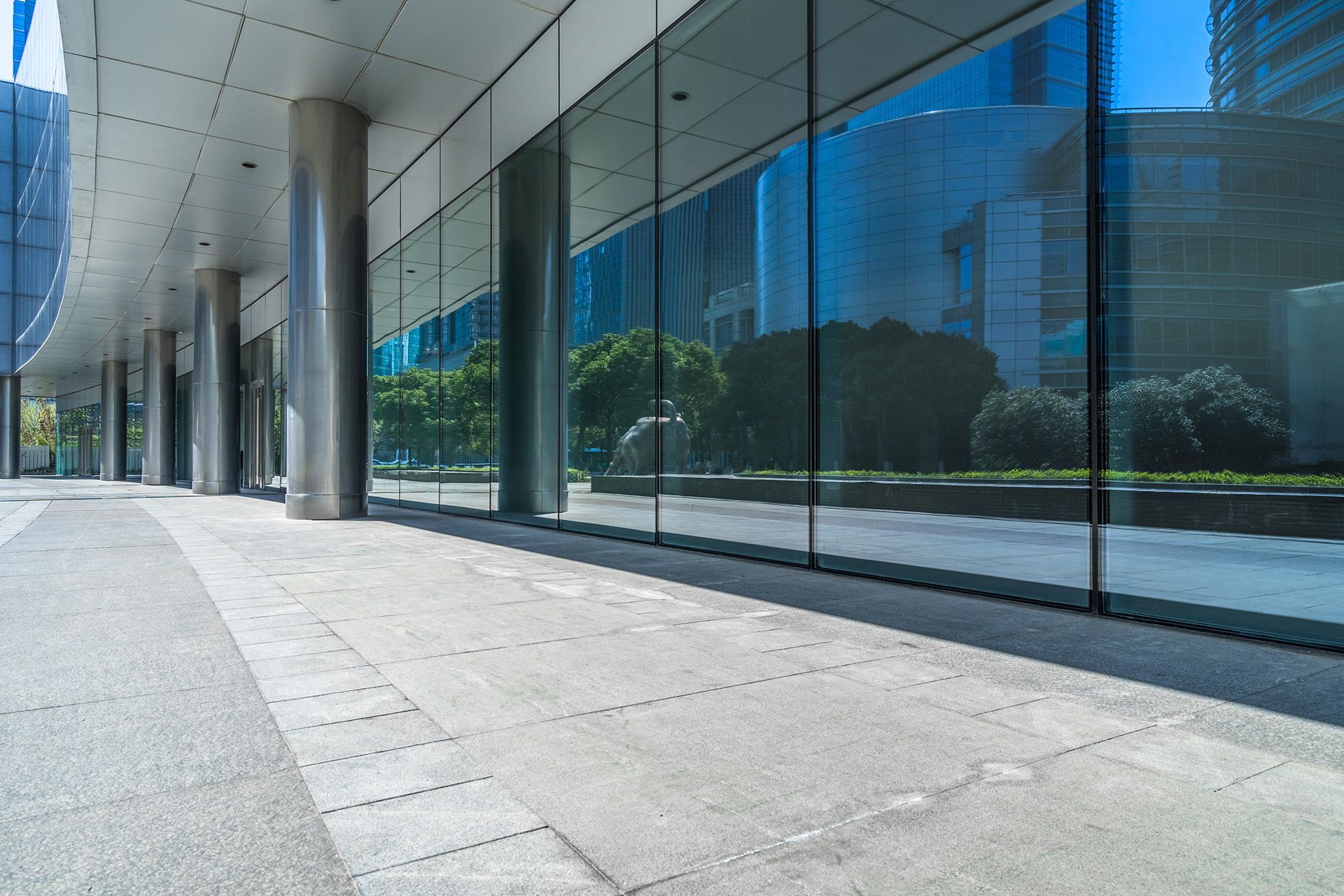 Modern building exterior with a glass facade reflecting the blue sky and surroundings. The sidewalk is empty with columns.