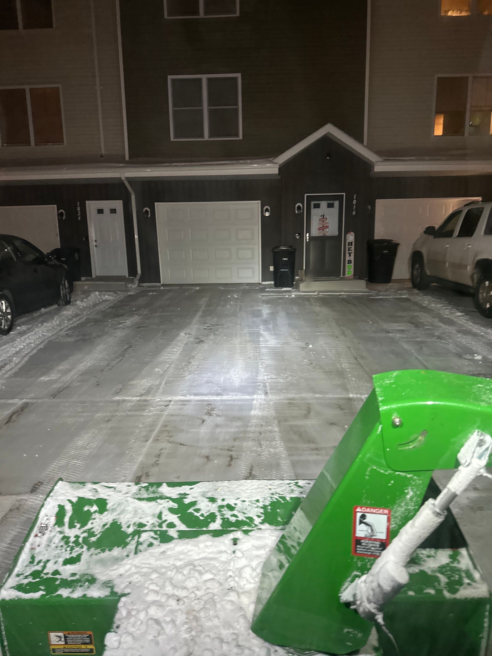 Green snowblower clearing snow from a driveway in front of an apartment building.