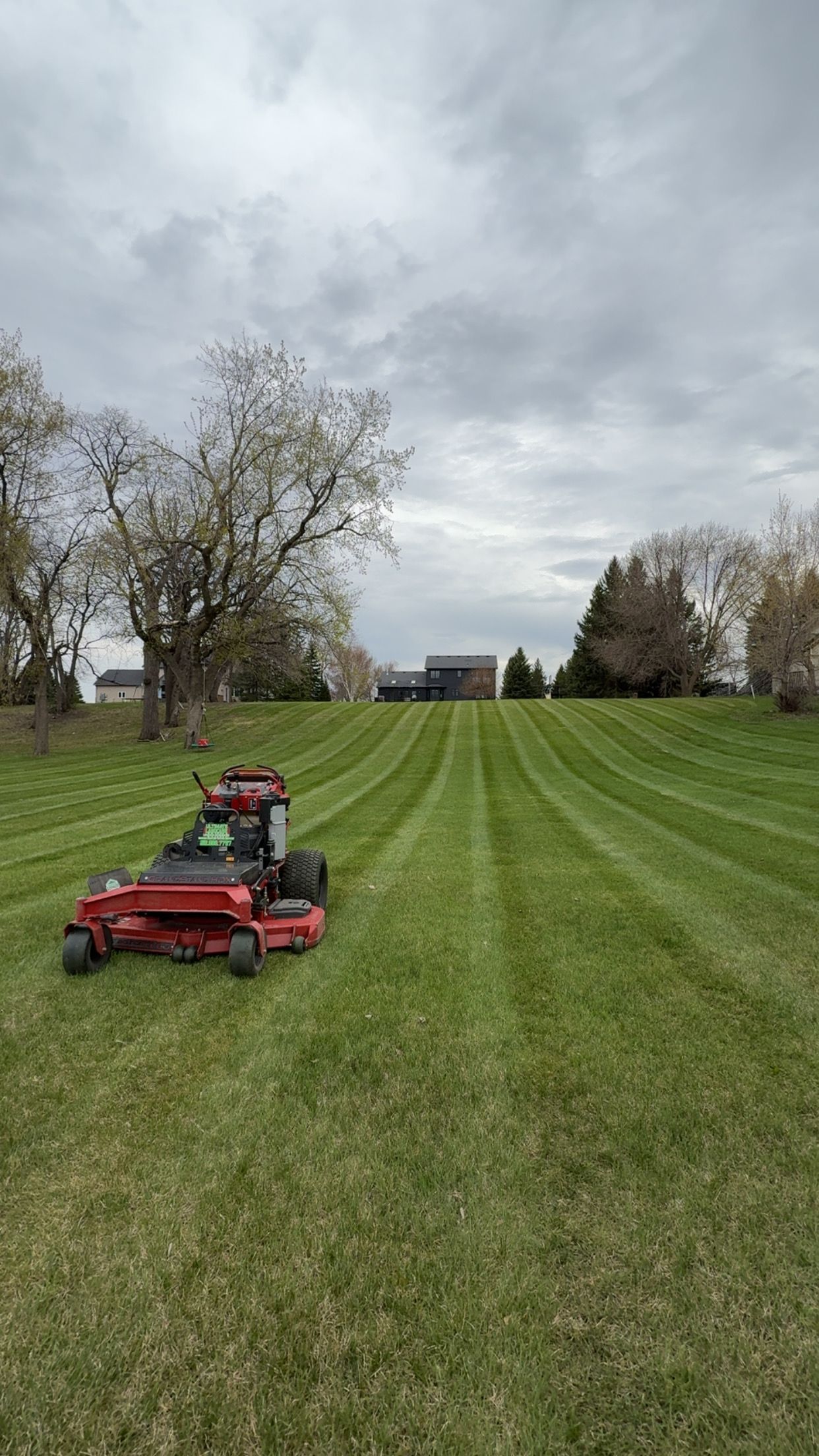 Red lawn mower on striped green field under cloudy sky, with trees and houses in the distance
