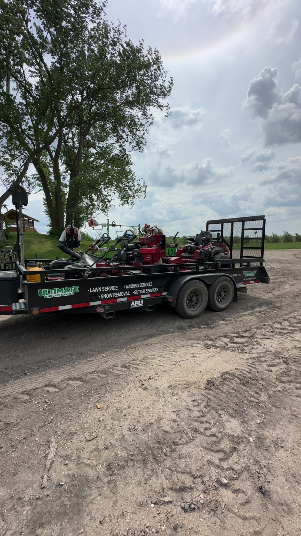 A black trailer carrying lawn equipment sits on gravel against a backdrop of a tree and cloudy sky.