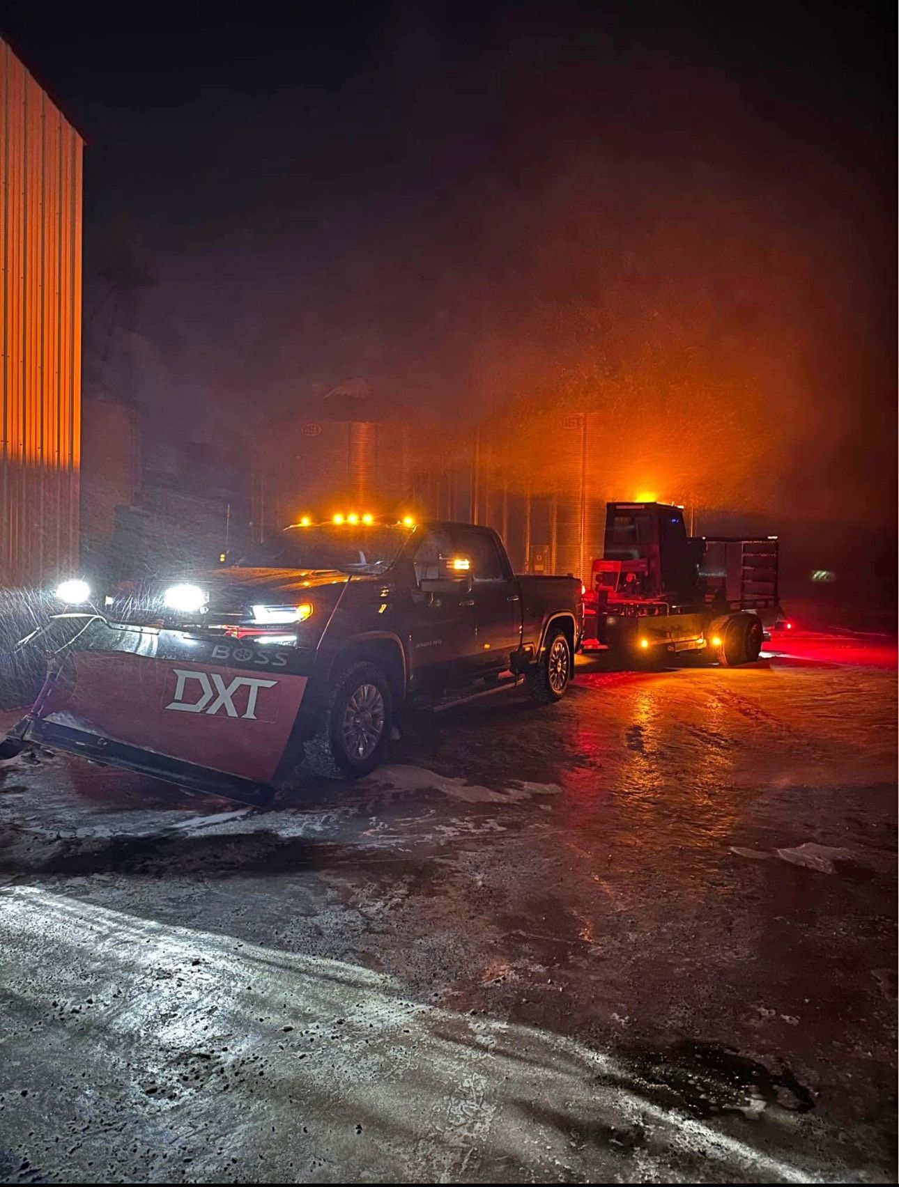 A dark pickup truck with a snowplow at night, towing a trailer.  Reflections on icy ground.  Orange and white lights.