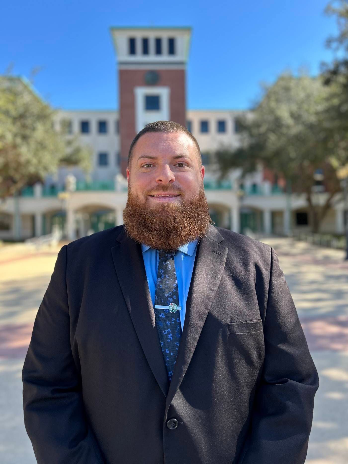 A man with a beard wearing a suit and tie is standing in front of a building.