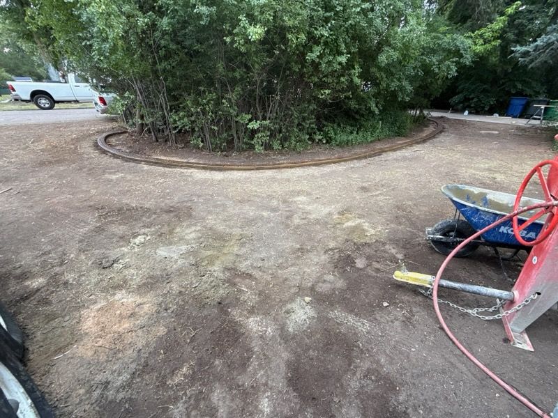 Dirt driveway with a curved brick border around a tree, wheelbarrow, and red equipment.