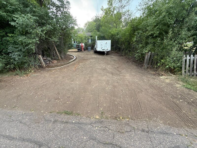 Dirt driveway leading to a white trailer and red tractor, flanked by trees and a wooden fence.