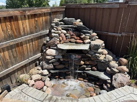 Stone water fountain cascading into a small pool, built against a wooden fence.