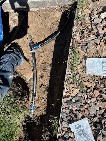 A trench with black tubing and connectors for an irrigation system, alongside gravel and grass; a person's leg is visible.
