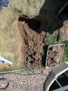 Hole dug in grass next to gravel and a window well. Soil pile nearby in a tray.
