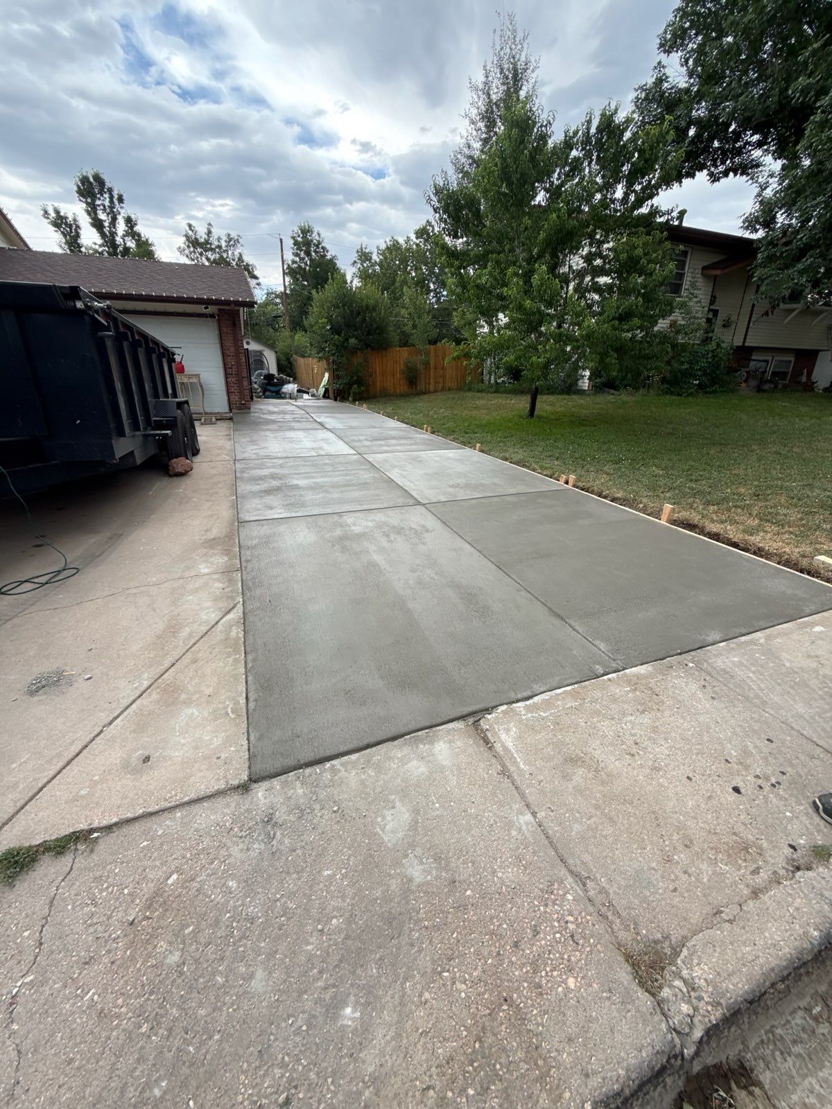 New concrete driveway section next to old sidewalk. A dark truck sits on the left, with trees and a house in the background.