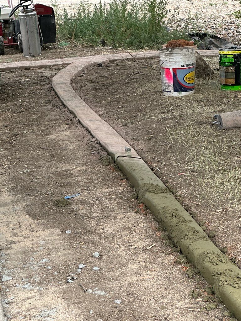 Concrete curb being installed outdoors in a dirt area; a bucket and tools are nearby.