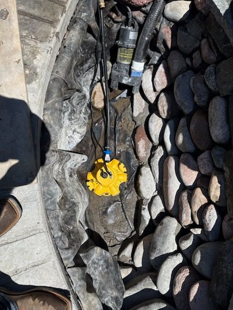 Yellow submersible pump in a rock-lined pond. Black hoses and electrical cords are connected.