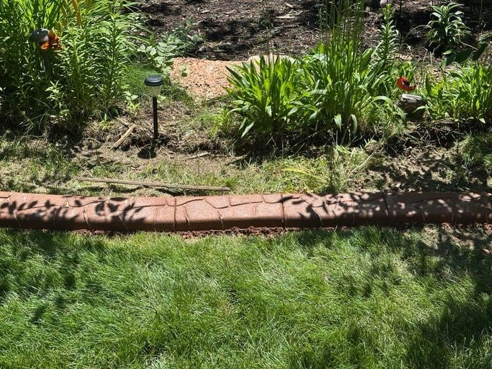 Lawn with a brick-like border separating green grass and a garden with plants, a solar light, and dark soil.