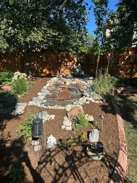Backyard with a cascading water feature, rocks, plants, and fresh mulch; trees in the background under a blue sky.