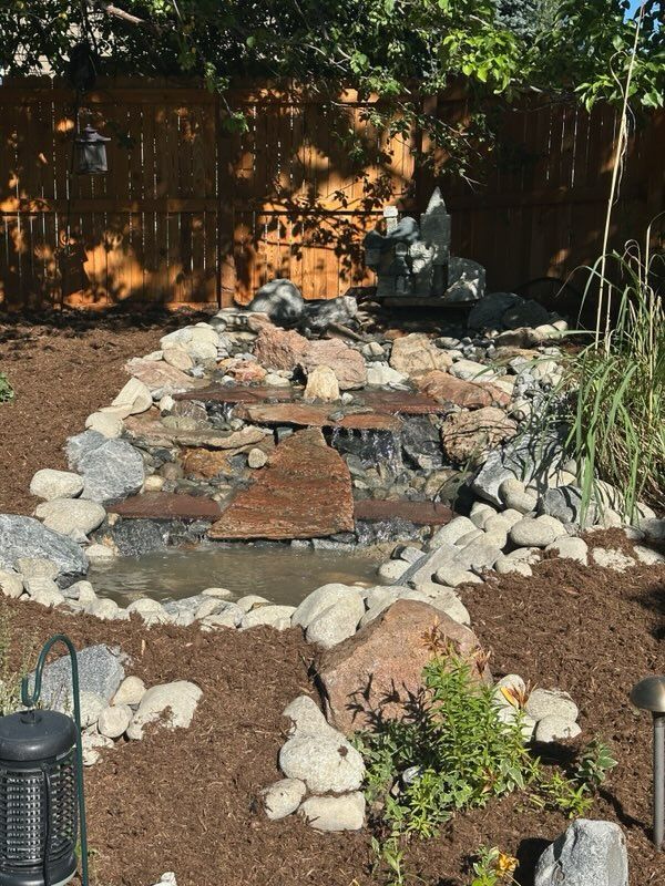 Backyard water feature with a waterfall, rocks, and mulch in front of a wooden fence.