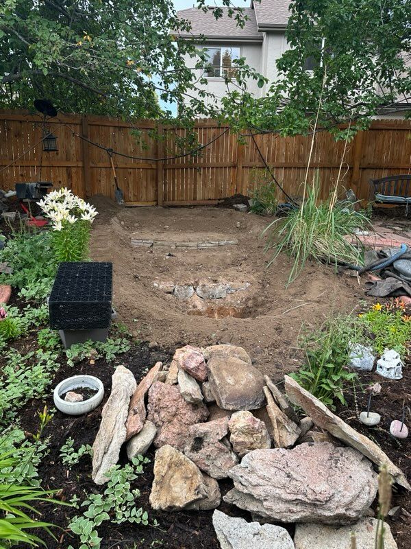Backyard with a dug-out area, surrounded by plants, rocks, and a wooden fence.