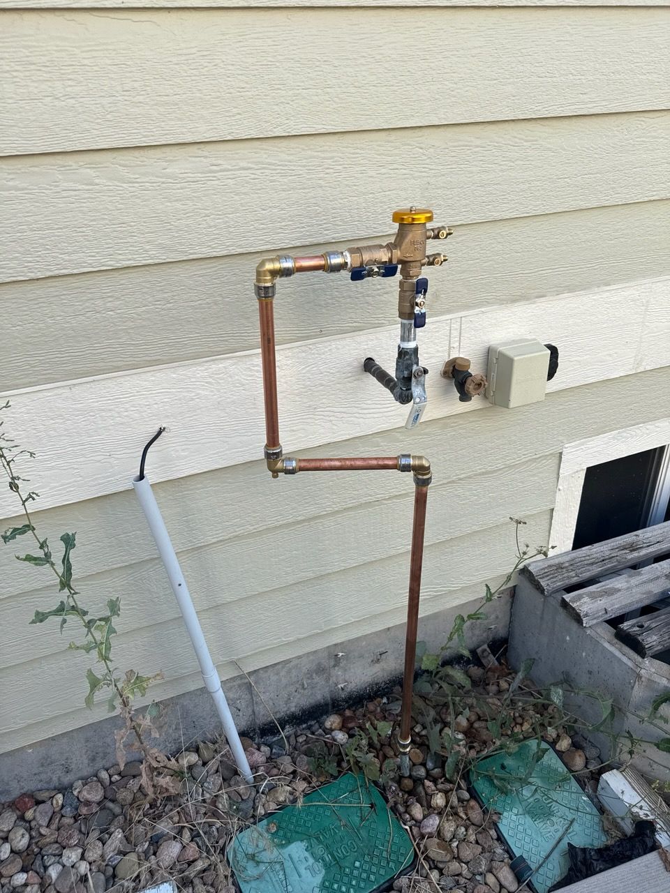 Copper plumbing pipes connected to a yellow-topped valve on a beige siding exterior wall.