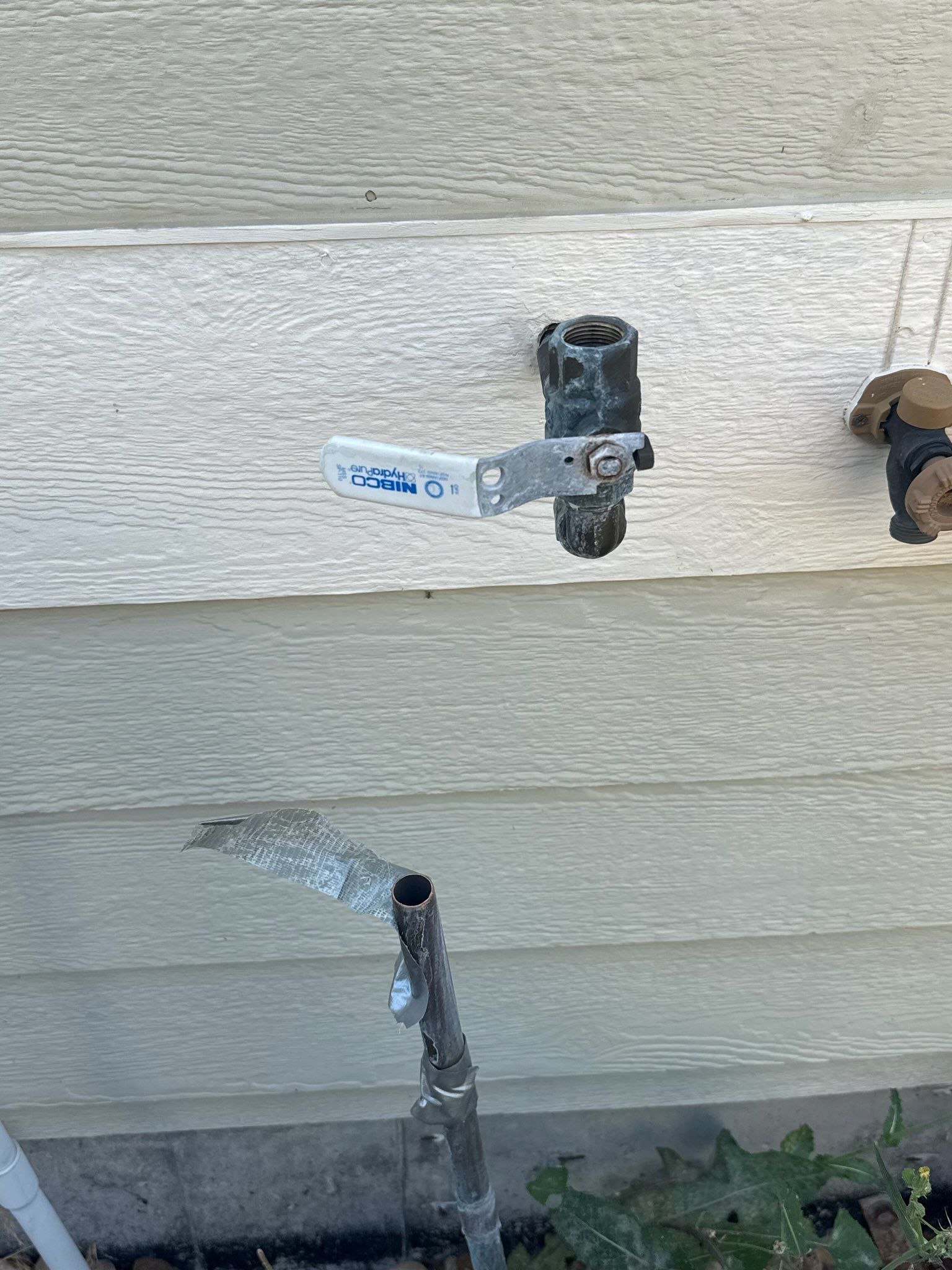 Water spraying from a broken pipe connected to an outdoor spigot on a beige house wall.