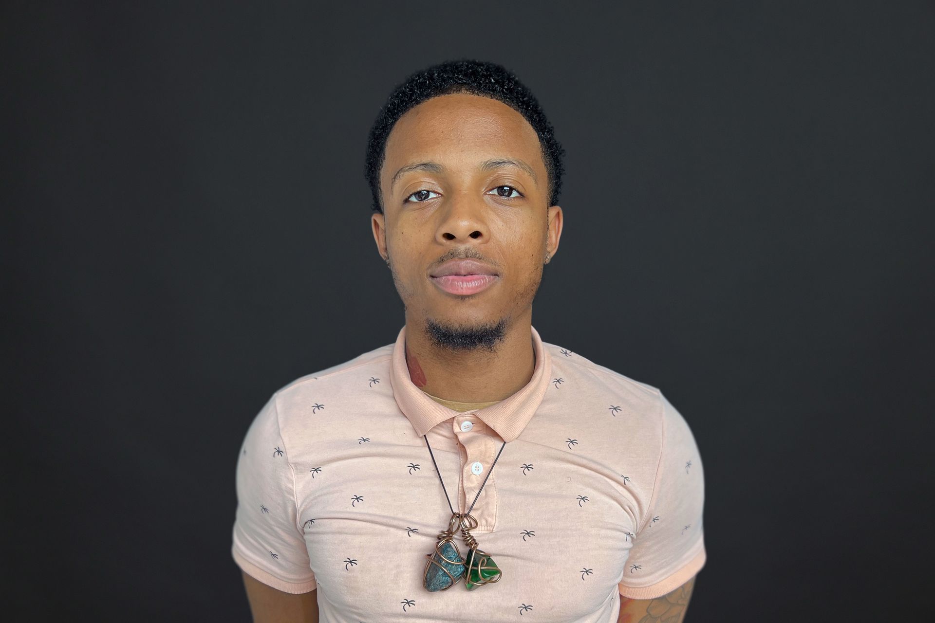 A young man wearing a pink polo shirt and a necklace is standing in front of a black background.
