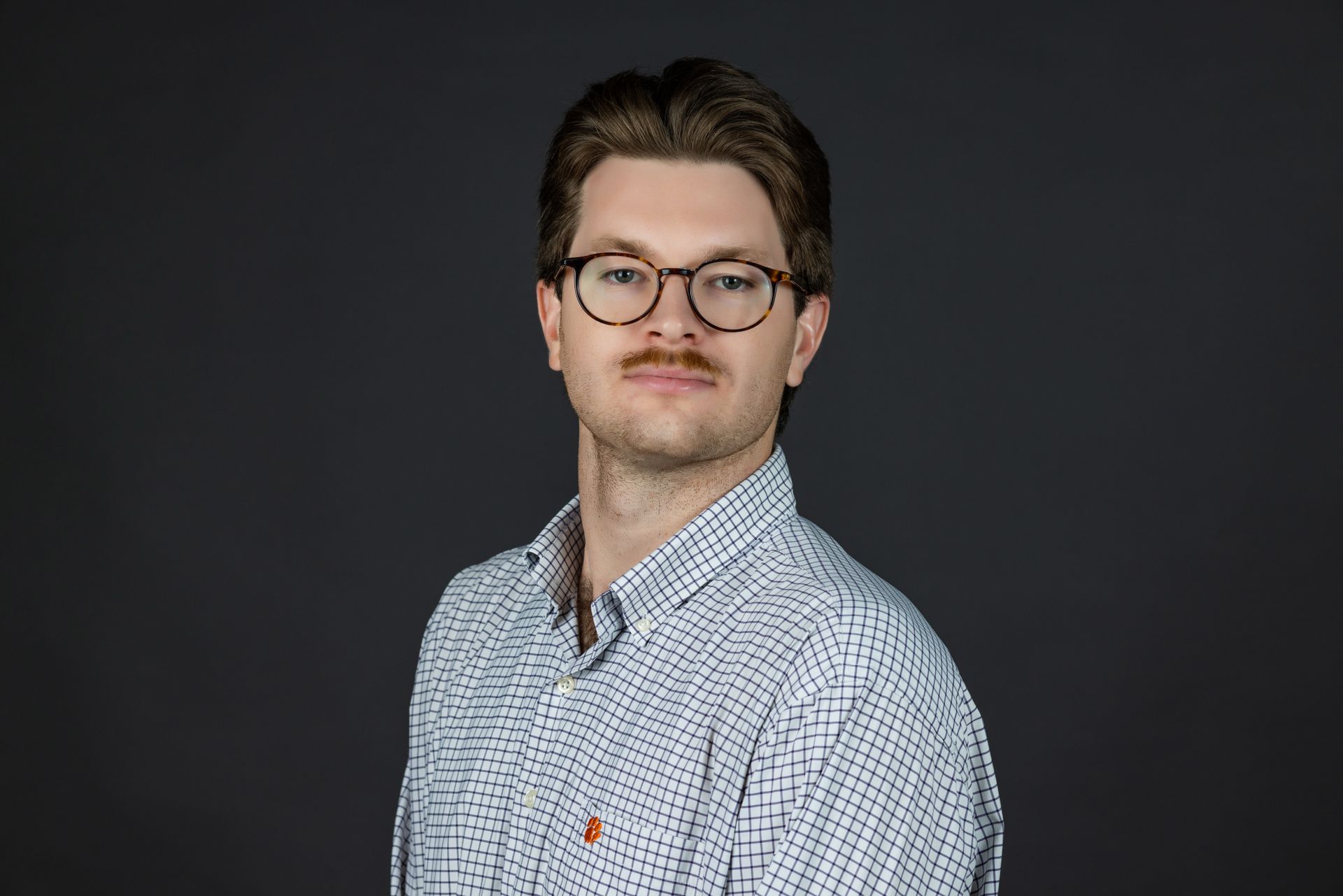 A young man wearing glasses and a plaid shirt is standing in front of a black background.