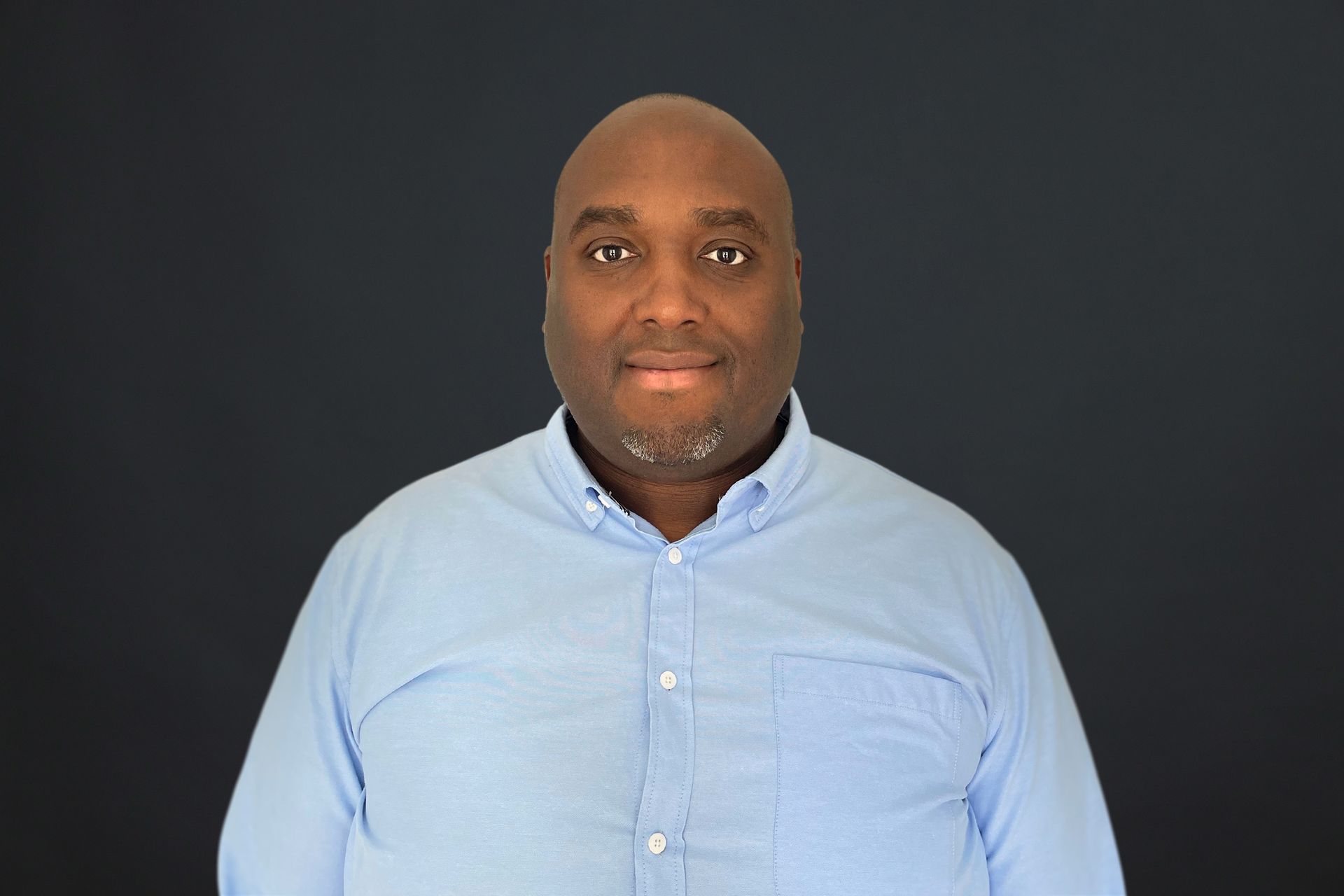 A man in a blue shirt is standing in front of a black background.