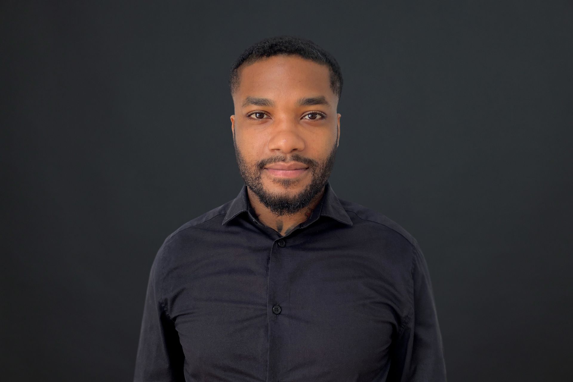 A man with a beard is wearing a black shirt and standing in front of a black background.