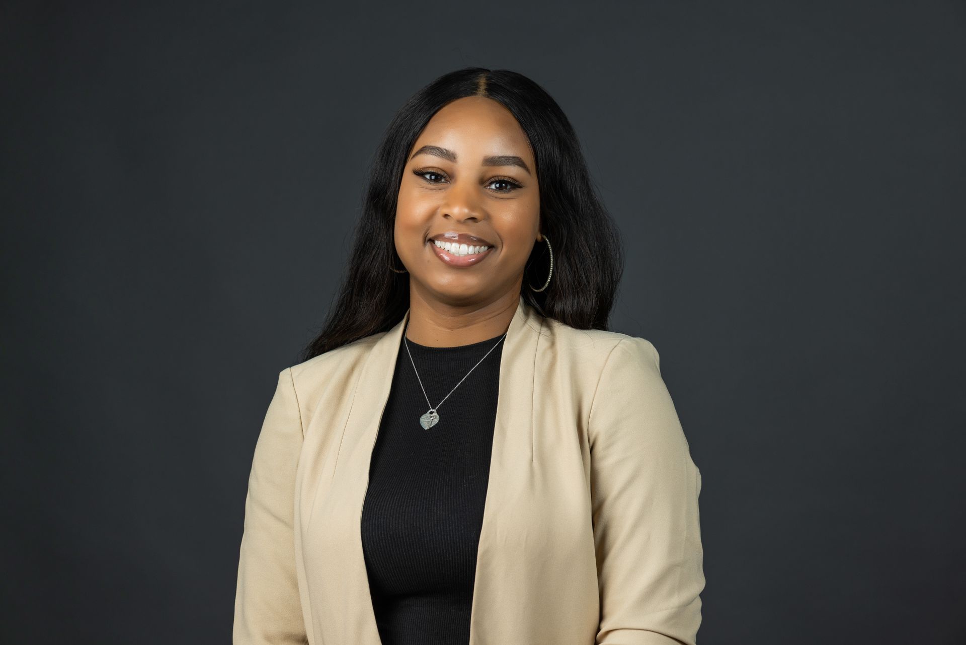A woman wearing a tan jacket and a black shirt is smiling for the camera.