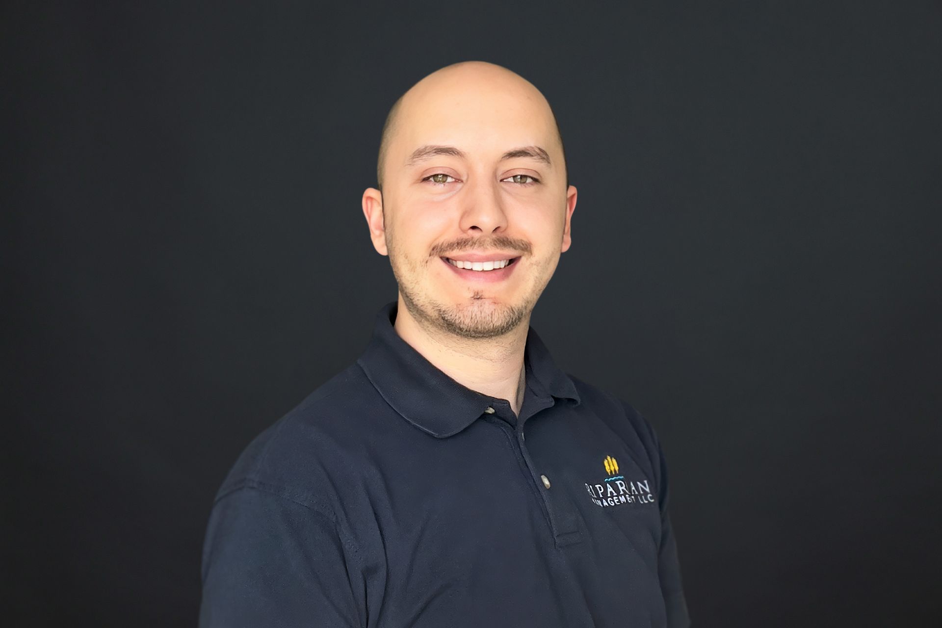 A bald man with a beard is wearing a blue polo shirt and smiling.