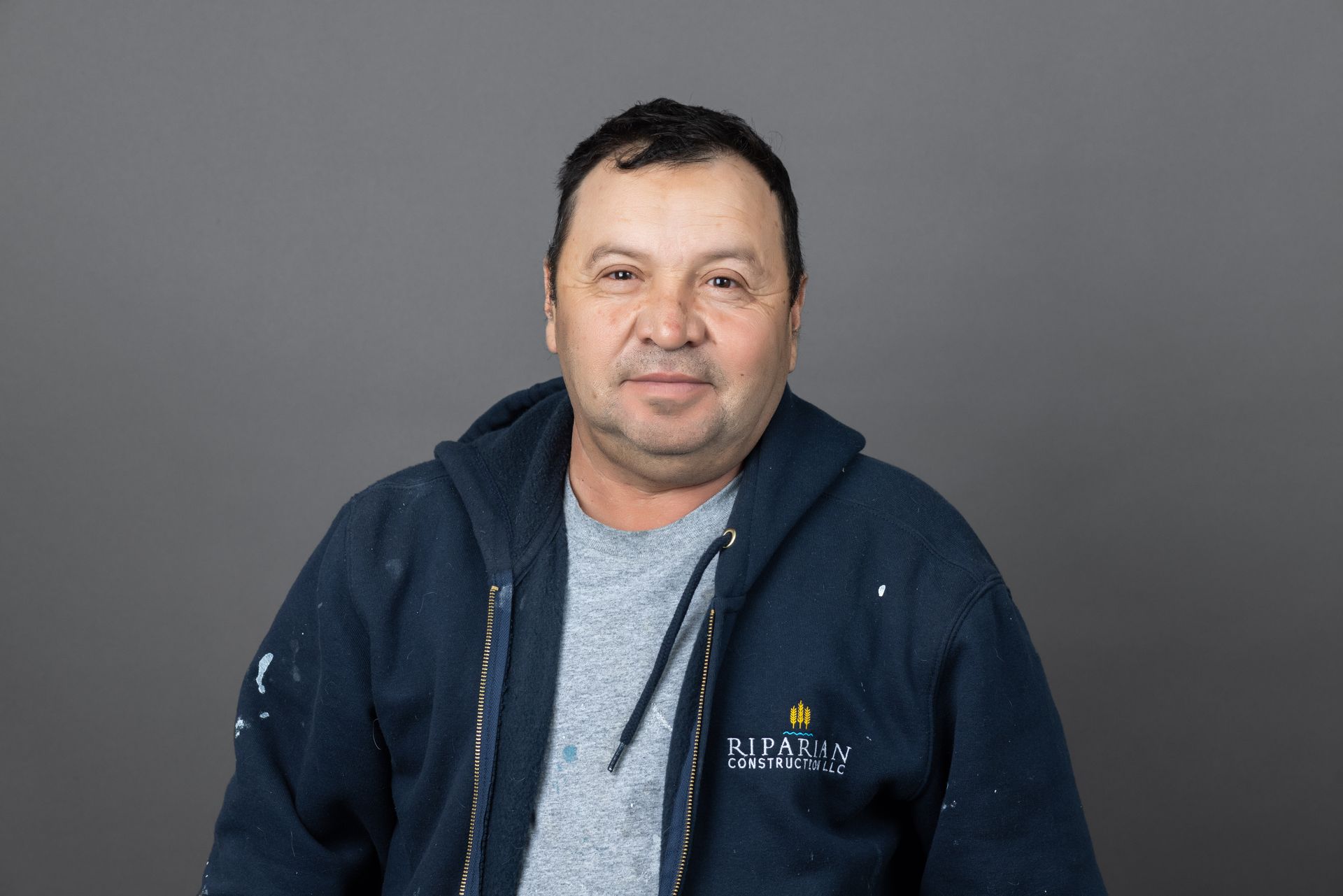 A man wearing a blue jacket and a grey shirt is standing in front of a gray background.