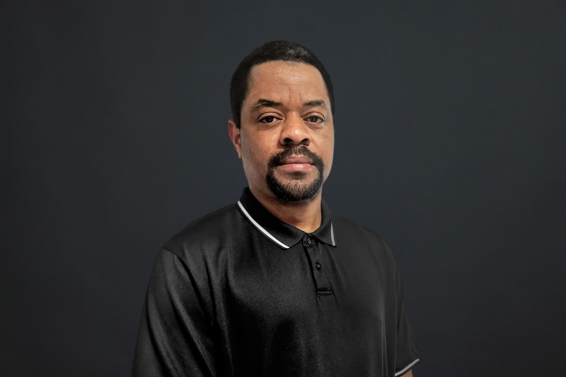 A man with a beard wearing a black polo shirt is standing in front of a black background.