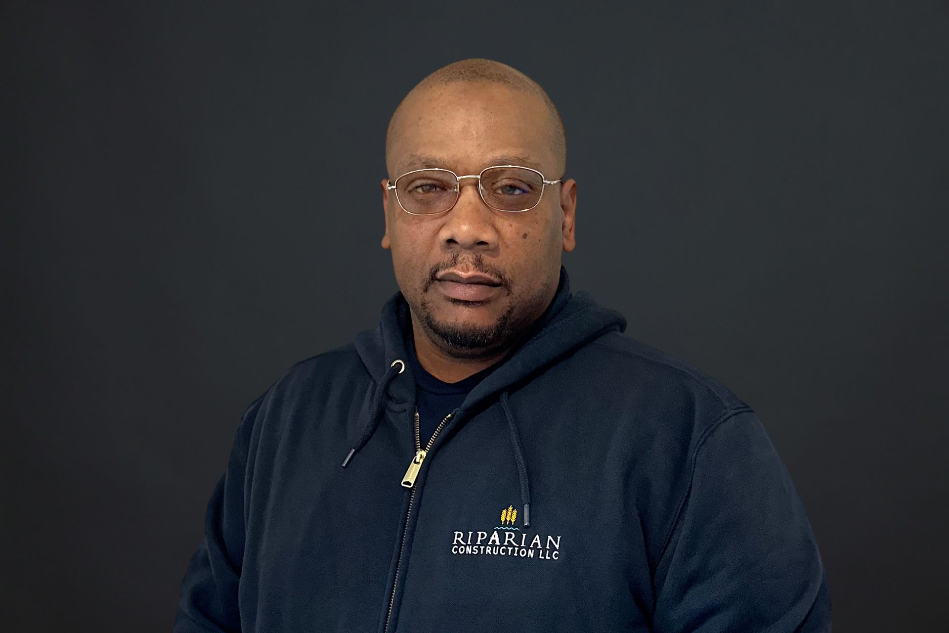 A man wearing glasses and a hoodie is standing in front of a black background.