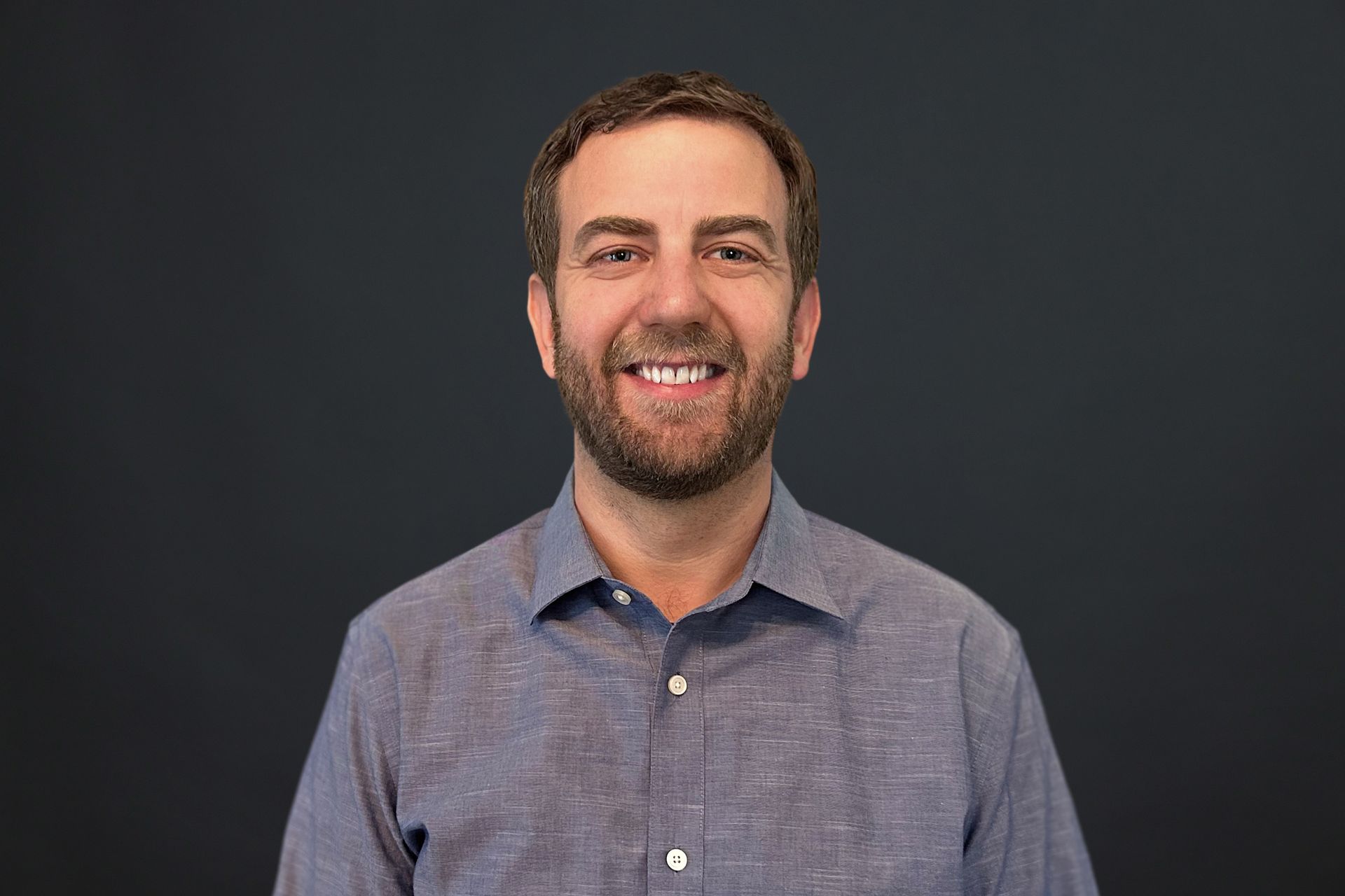 A man with a beard is smiling for the camera while wearing a blue shirt.