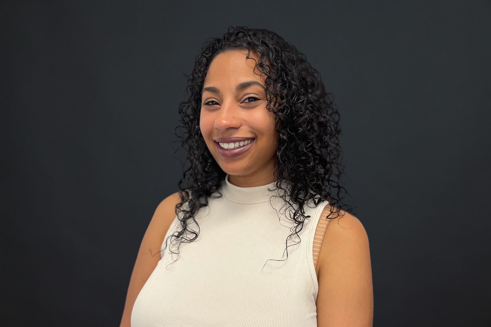 A woman with curly hair is wearing a white tank top and smiling.