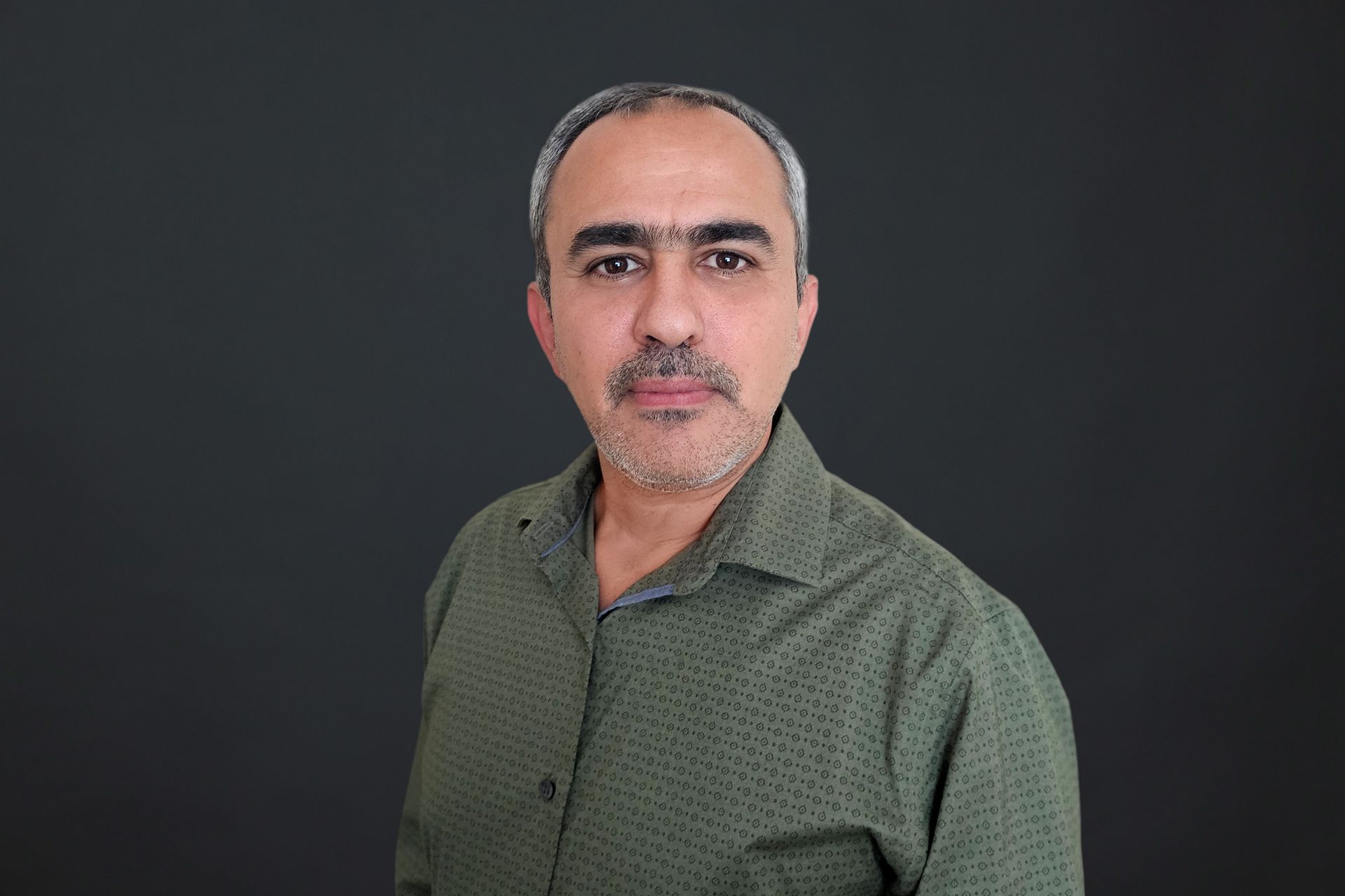 A man with a beard is wearing a green shirt and standing in front of a black background.