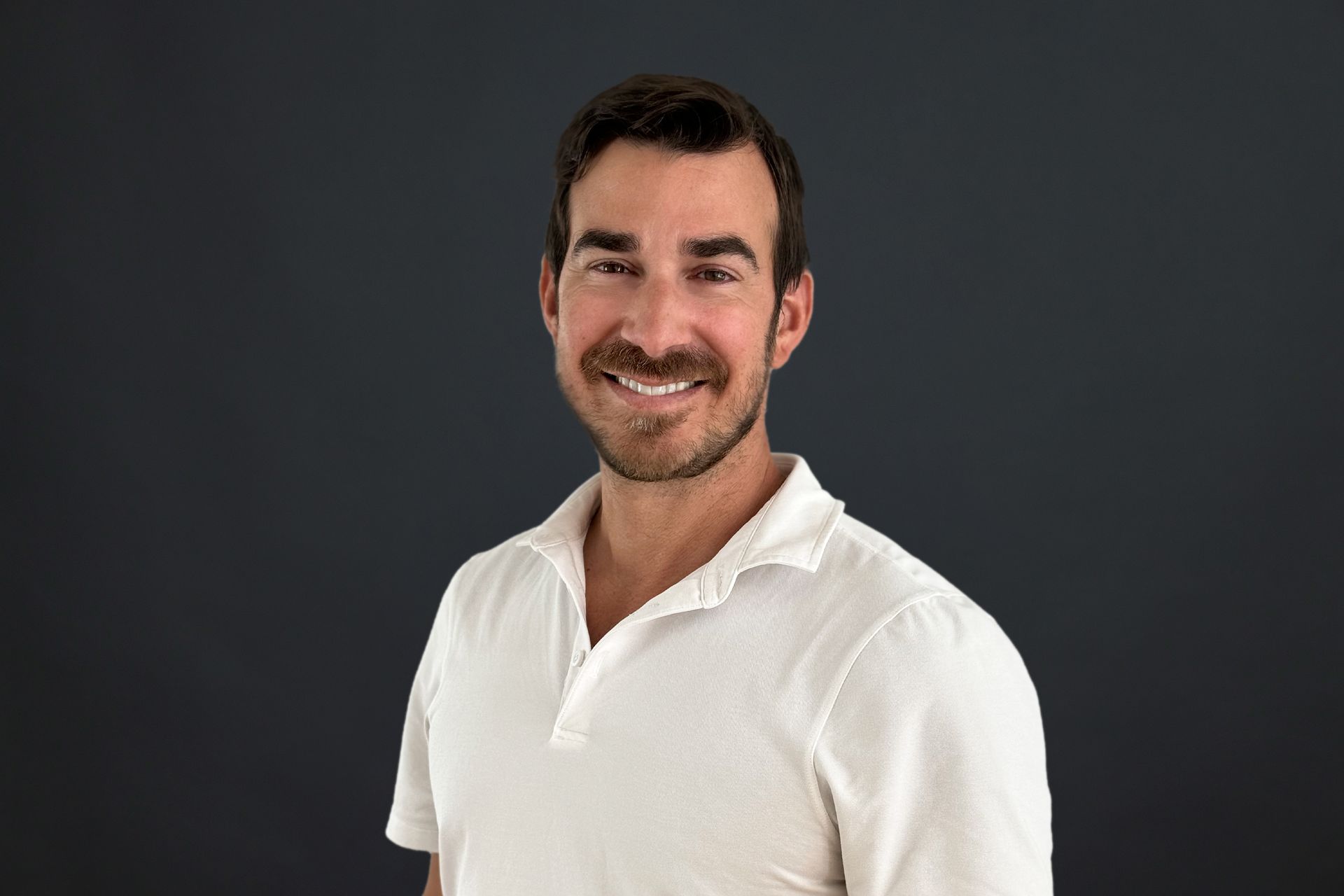 A man with a beard is wearing a white polo shirt and smiling.