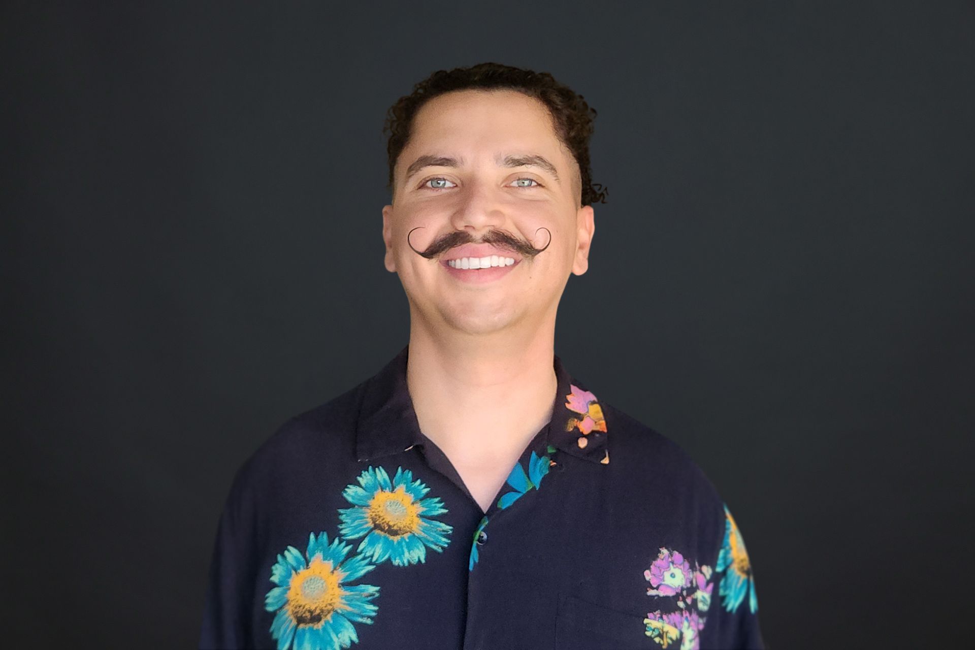 A man with a mustache is wearing a floral shirt and smiling.