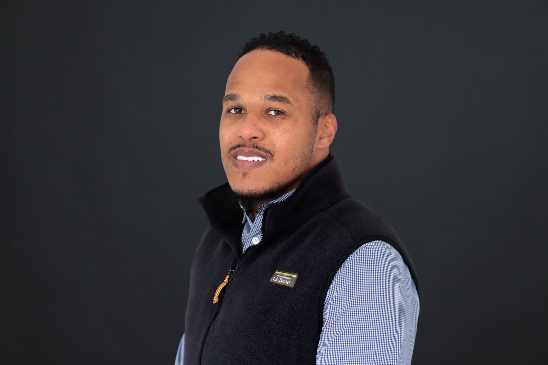 A man wearing a black vest and a plaid shirt is standing in front of a black background.