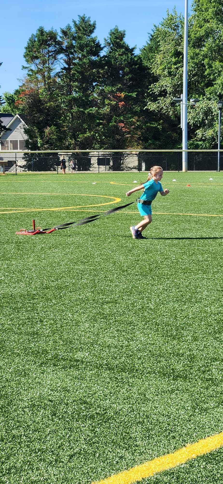 A person in a blue shirt and shorts runs across a bright green sports field, pulling a weighted sled with a black strap.