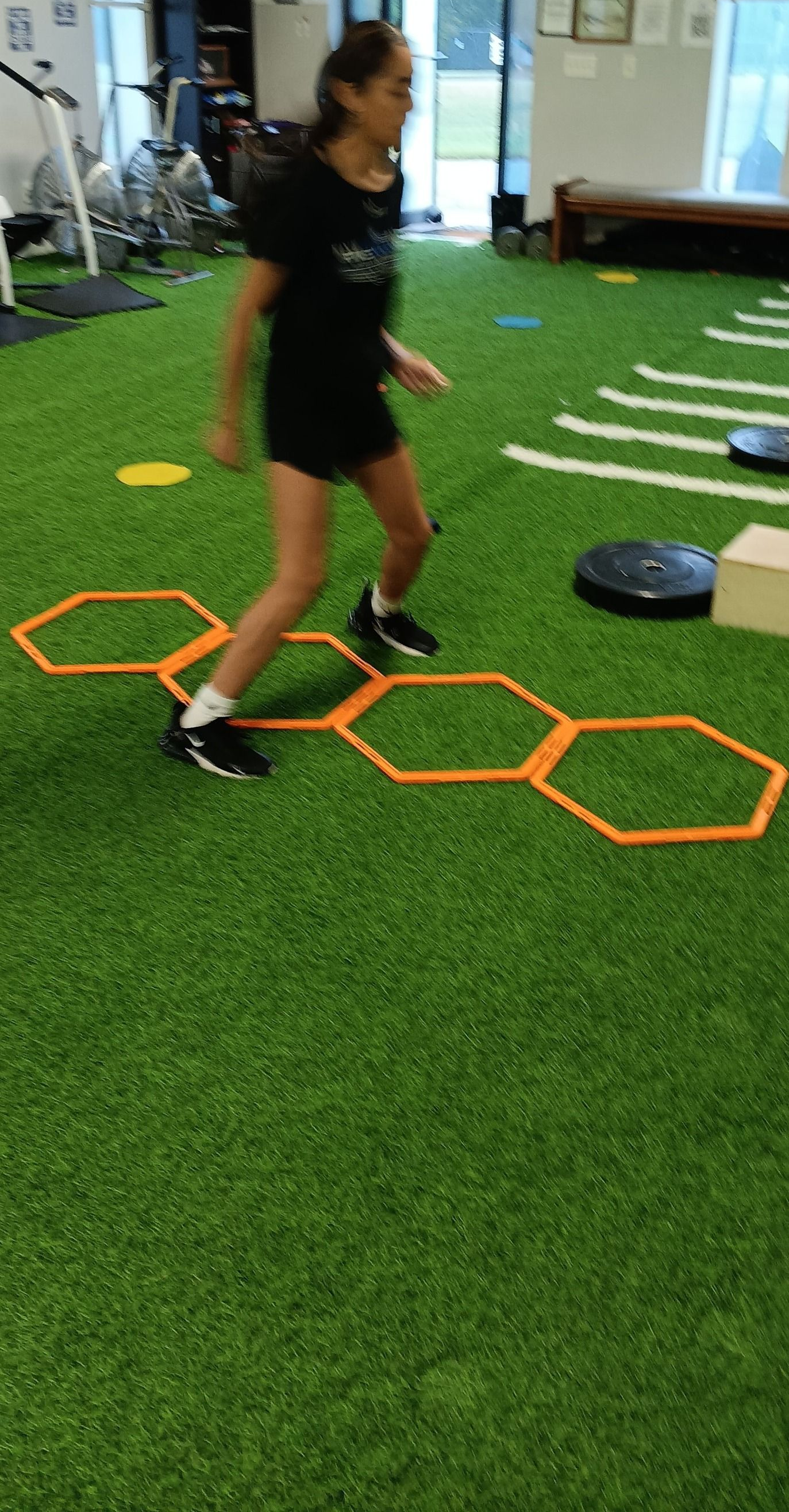 A person performs agility training using orange hexagon-shaped rings laid out on a green gym floor.
