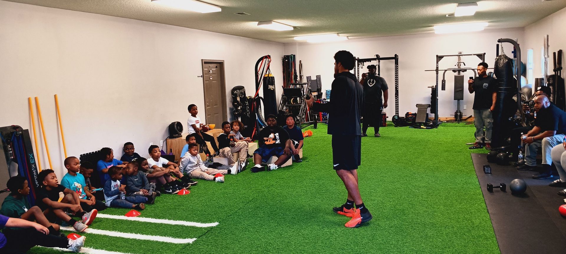 A coach speaks to a group of children sitting on a turf-covered indoor training floor with fitness equipment in the back.