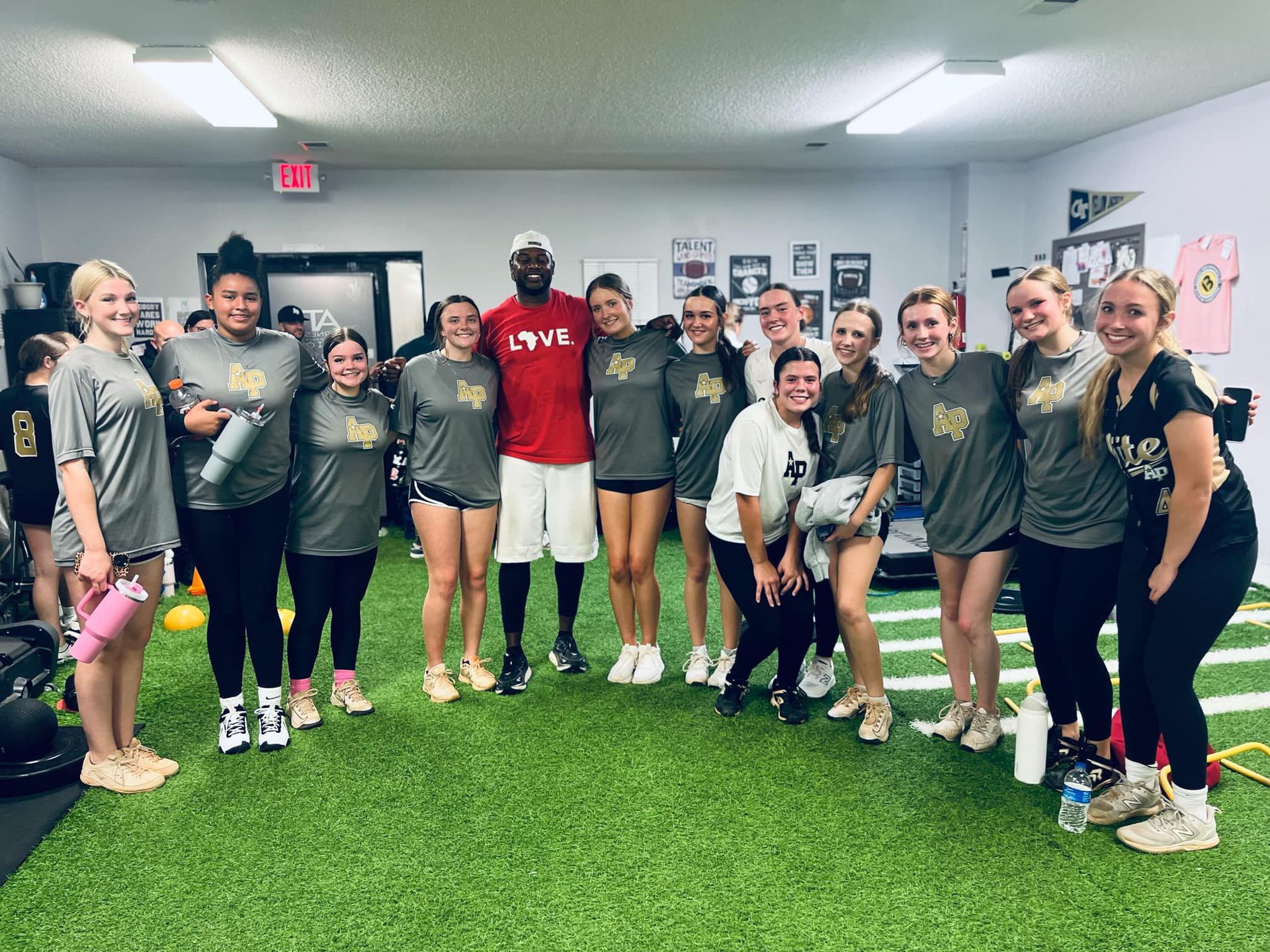 A group of people in athletic wear pose together on an indoor turf field with a coach in a red shirt.