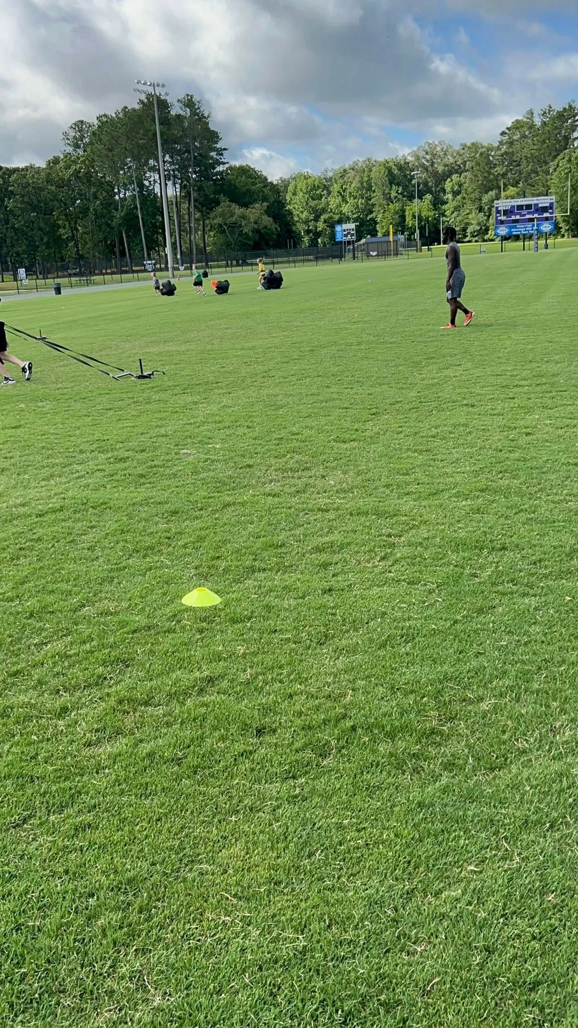 People exercise on a grassy field with yellow cones arranged on the ground under a cloudy sky.