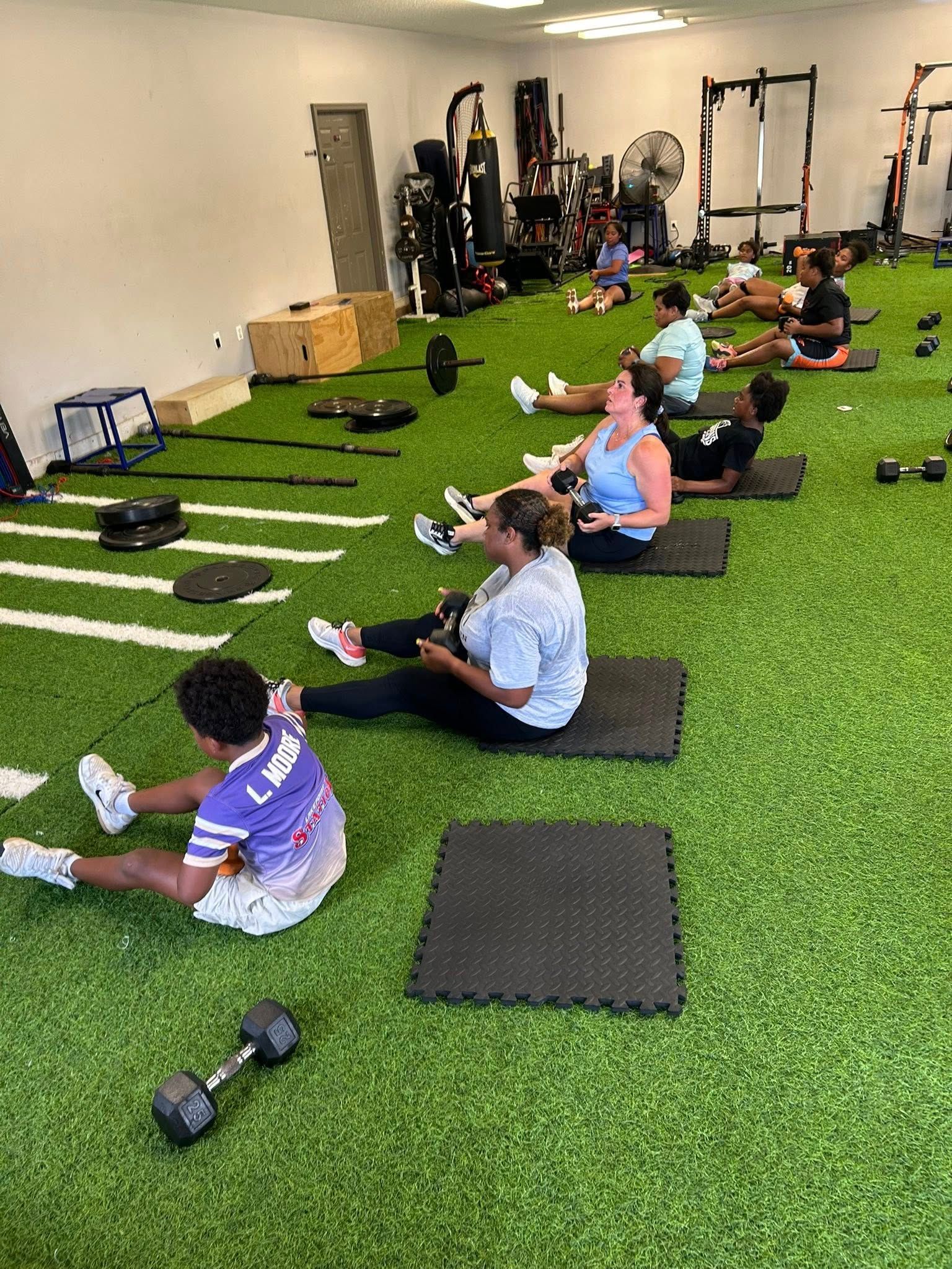 People sit on mats in a gym with artificial grass, performing core exercises with dumbbells.
