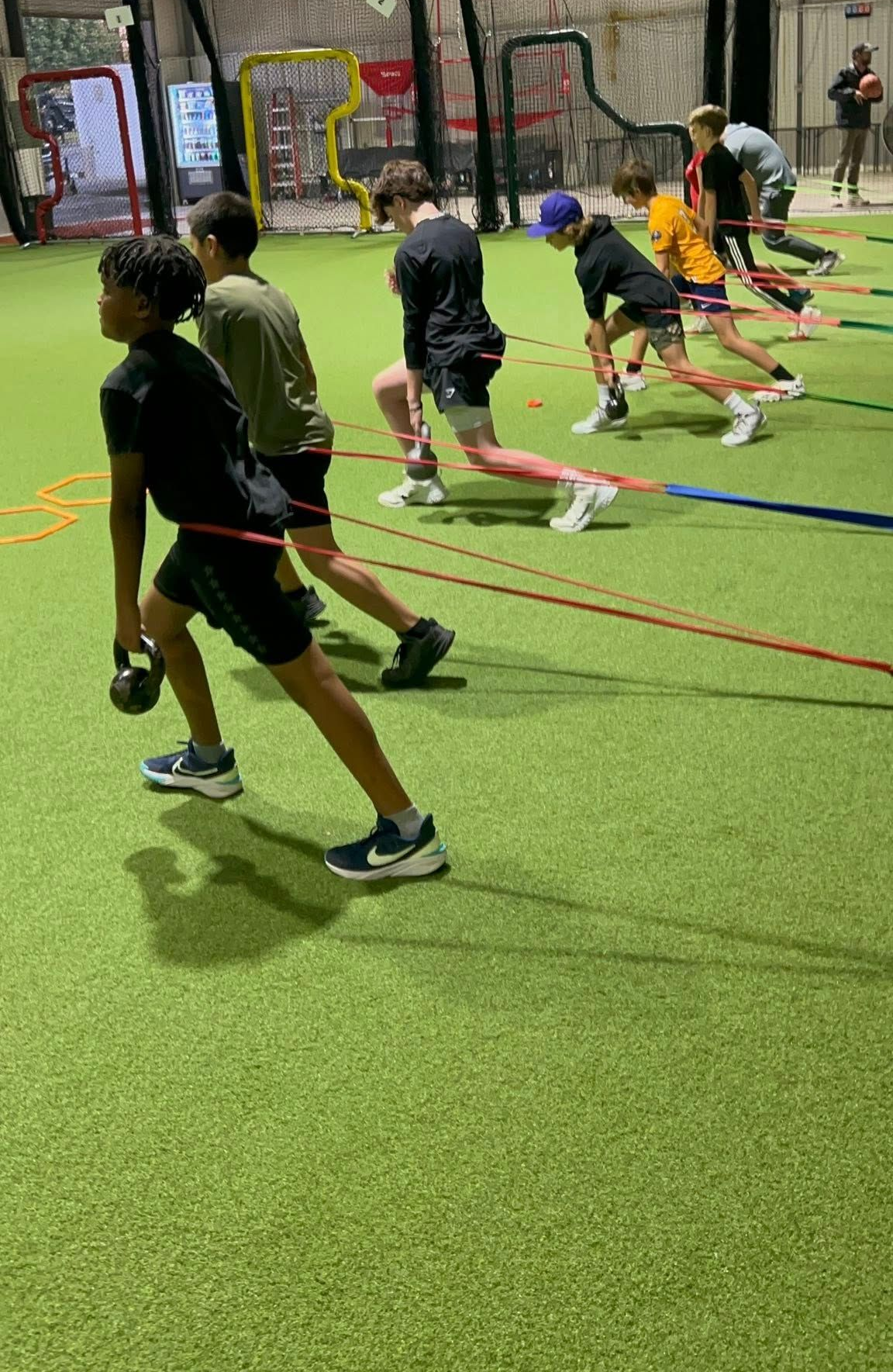 A row of people in athletic wear perform weighted lunges on an indoor turf field, holding kettlebells and resistance bands.