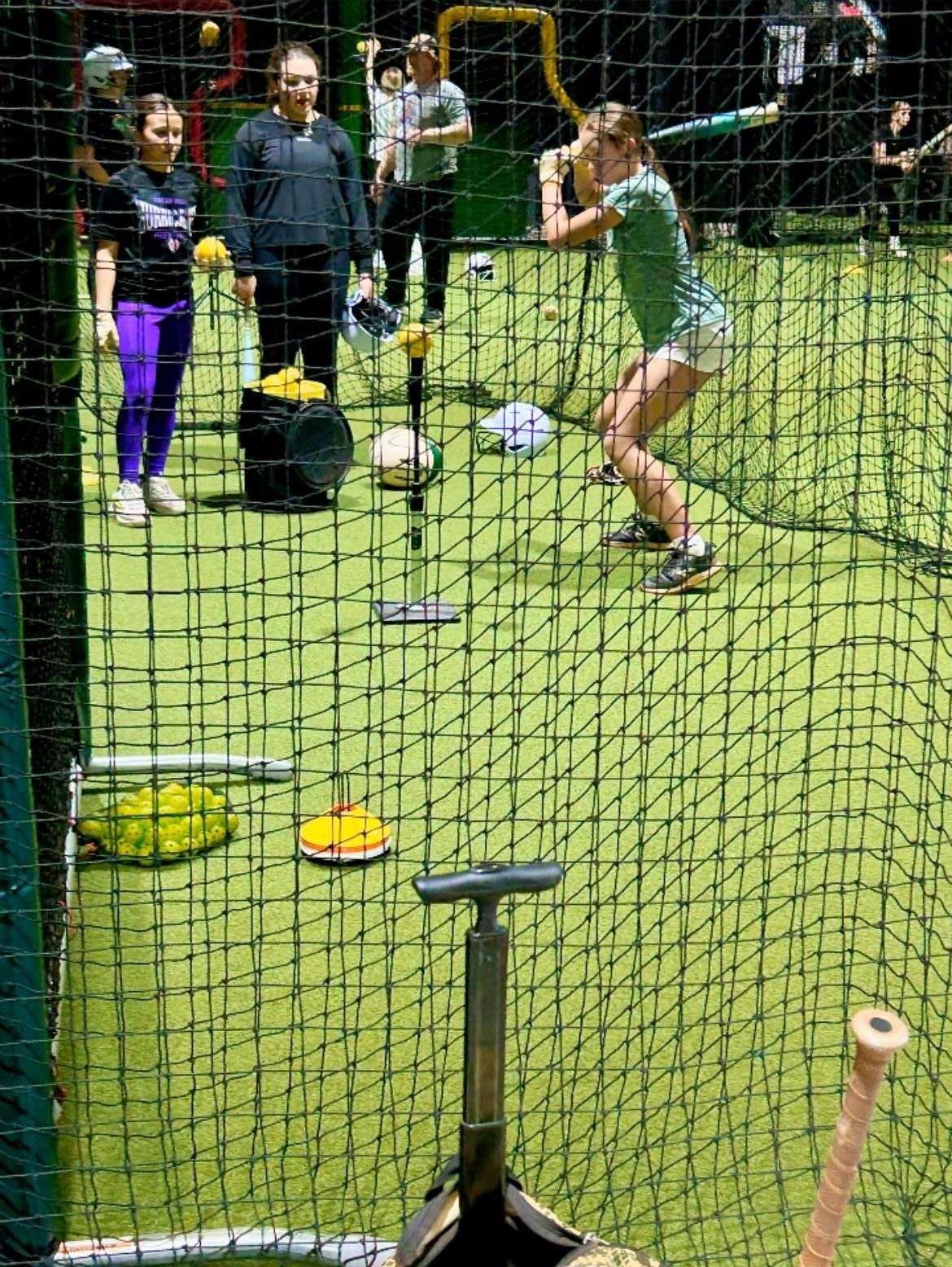 A batting practice session in an indoor cage, with a person swinging a bat at a ball on a tee as others watch nearby.